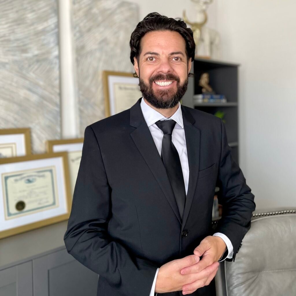 Attorney Chaz D, Lyons standing beside his diplomas, smiling warmly in a well-dressed suit.